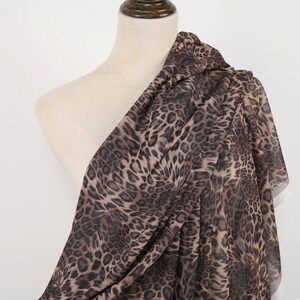 May include: A draped fabric with a leopard print pattern. The fabric is draped over a mannequin, showcasing the flowing material. The print features brown and black spots on a tan background. The fabric appears lightweight and semi-sheer.