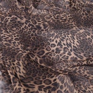 May include: Close-up of a fabric with a leopard print pattern. The fabric features a tan background with dark brown and black spots. The material is draped, showing folds and texture.