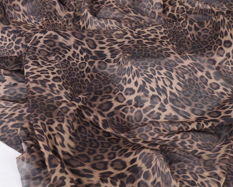 May include: Close-up of a fabric with a leopard print pattern. The fabric features a tan background with dark brown and black spots. The material is draped, showing folds and texture.
