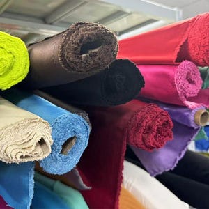 May include: A variety of fabric rolls in different colors and textures, including green, yellow, brown, black, red, pink, purple, blue, and beige. The rolls are stacked on top of each other and are arranged in a row.