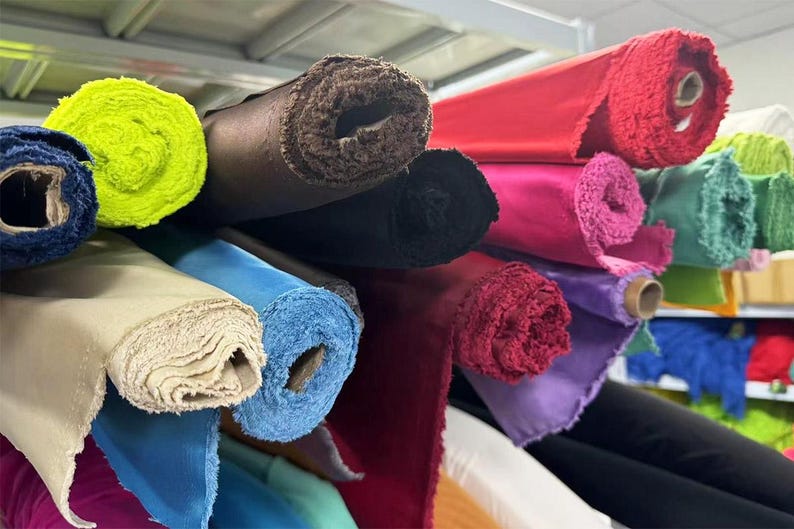 May include: A variety of fabric rolls in different colors and textures, including green, yellow, brown, black, red, pink, purple, blue, and beige. The rolls are stacked on top of each other and are arranged in a row.