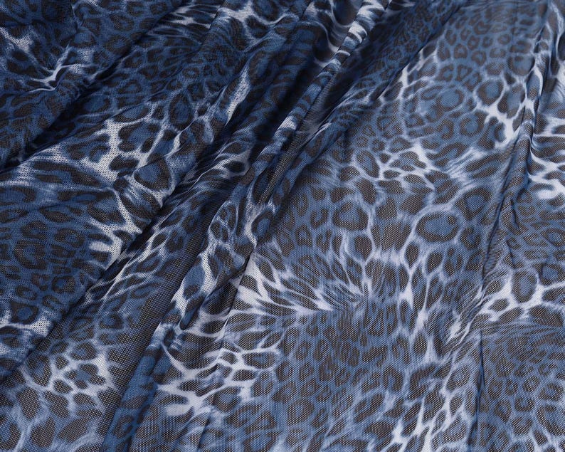 May include: Close-up of a blue and white leopard print fabric. The fabric has a sheer, mesh-like texture with a pattern of dark blue and white spots. The fabric is draped, creating folds and shadows.