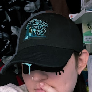 Może przedstawiać: Czarna czapka z daszkiem z niebieską i turkusową naszywką. Czapka ma zakrzywiony daszek i siatkową tylną część. Osoba na zdjęciu ma na sobie szarą koszulkę z długim rękawem.