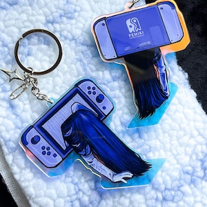 Puede incluir: Dos llaveros con un diseño de consola de videojuegos azul y una figura de pelo largo. Uno tiene un charm de estrella y un imperdible. El otro tiene la palabra "FEMIKI" impresa. Ambos tienen un llavero plateado.