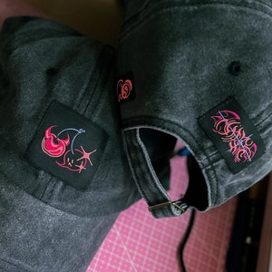 May include: Two black denim baseball caps with embroidered patches. One patch features a red and pink cherry with a star, the other a red and pink flame design.