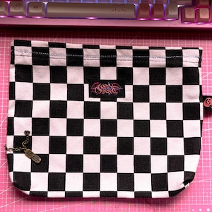 May include: A black and white checkered pouch with a small metal charm attached. The pouch has a black and white checkered pattern and a small black tag with the word "Grunge" on it.