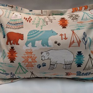 Pode incluir: Uma fronha bege com um padrão repetido de ursos, tipis, flechas e desenhos geométricos em tons de azul, laranja e marrom. O texto "Bear" aparece duas vezes na fronha.