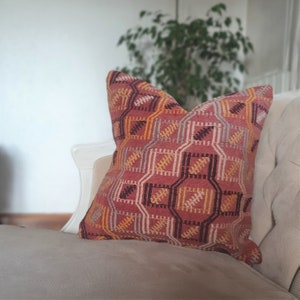 Puede incluir: Una almohada de tiro con estampado rojo, naranja, amarillo y marrón con un diseño geométrico. La almohada está sobre un sofá beige claro.