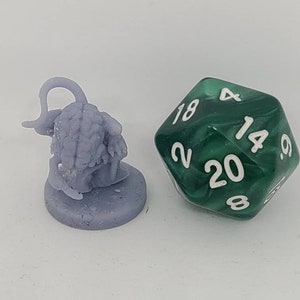 May include: A small, purple, plastic miniature of a creature with a curled tail and a textured body. The miniature is on a round base. A green 20-sided die with white numbers is in the background.