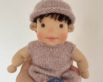 May include: A knitted doll with a pink hat and outfit, blue knitted shoes, and brown hair. The doll has a light brown complexion and painted features.
