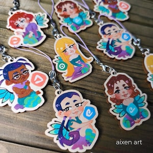 May include: Wooden charm pendants featuring colorful illustrations of fairy-like characters with wings. Each charm has a unique design, with variations in hair color, clothing, and accessories. The charms are attached to a silver-colored clasp and a purple lanyard.