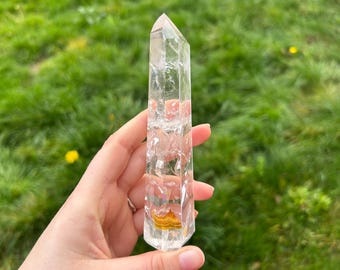 May include: A clear quartz crystal point with a faceted shape. The crystal has internal inclusions and a golden-brown mineral deposit at the base. The background is blurred green grass.