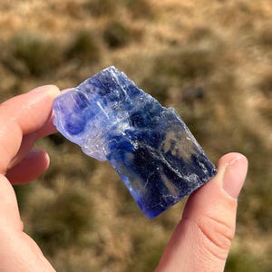 May include: A raw, rectangular crystal specimen featuring a gradient of translucent light blue to deep navy blue coloration. The mineral has a jagged, uneven surface texture with visible internal fractures and crystalline layers throughout the stone.