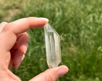 Puede incluir: Una punta de cristal de cuarzo transparente, sostenida entre los dedos sobre un fondo verde borroso. El cristal es translúcido con estructuras internas visibles y una punta facetada, mostrando la belleza natural.