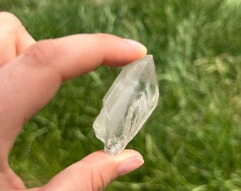 Puede incluir: Un cristal de cuarzo claro y facetado, sostenido entre los dedos, sobre un fondo verde borroso. El cristal es translúcido con estructuras internas visibles y una punta afilada.
