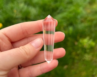 May include: A clear crystal point with a faceted design. The top is a reddish-pink color, with bands of green and clear sections along the length. The crystal is held in a hand against a blurred green background.