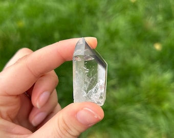 Puede incluir: Una punta de cristal de cuarzo transparente, sostenida entre los dedos, con una forma natural y facetada. El cristal es transparente con fracturas e inclusiones internas. El fondo es hierba verde borrosa.