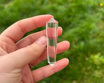 May include: A clear, faceted crystal wand held in a hand against a blurred green background. The crystal is rectangular with a pointed tip and a flat top. The facets create a striped effect within the crystal.
