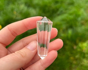 May include: A clear quartz crystal point, held in a hand, with a faceted top and a tapered, ridged body. The crystal is transparent, allowing light to pass through. The background is blurred green foliage.