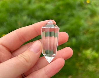 May include: A clear, faceted crystal point with a pointed tip. The crystal is held against a blurred green background of grass. The crystal is approximately 3 inches long.