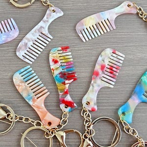 May include: A collection of colorful comb keychains. Each comb features a unique marbled pattern in shades of pink, blue, green, and red. The keychains have gold-colored hardware, including a clasp and a keyring.