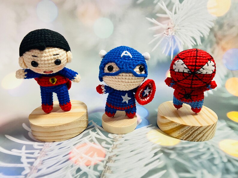 May include: Three crocheted superhero figurines: Superman, Captain America, and Spiderman. Each figurine is standing on a wooden base.