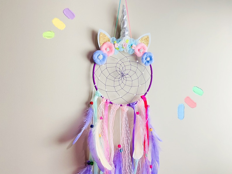 K&ouml;nnte beinhalten: Ein lila und wei&szlig;er Traumf&auml;nger mit einem Einhornhorn und Blumenschmuck. Der Traumf&auml;nger hat Federn und Perlen, die von ihm herabh&auml;ngen.
