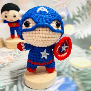 May include: A crocheted Captain America doll with a red, white, and blue costume, holding a red, white, and blue shield. The doll is standing on a wooden base.