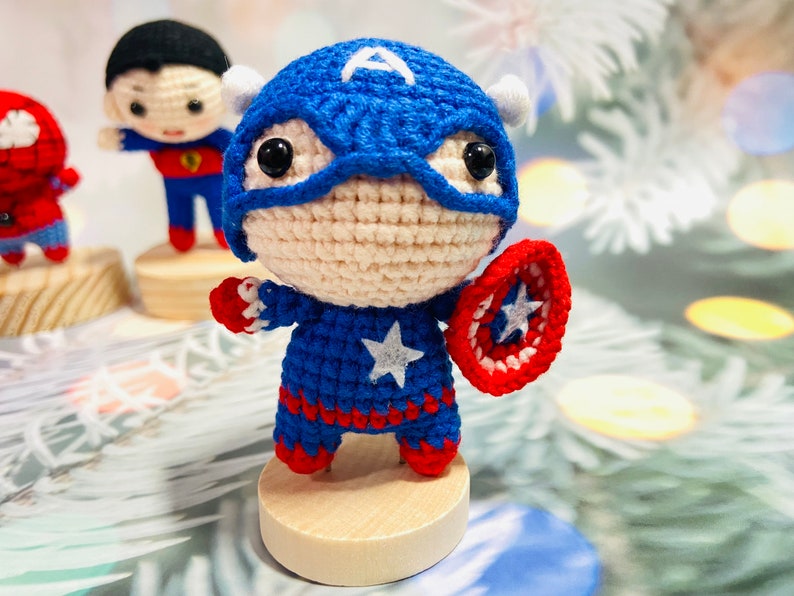 May include: A crocheted Captain America doll with a red, white, and blue costume, holding a red, white, and blue shield. The doll is standing on a wooden base.