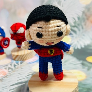 May include: A crocheted superhero doll with a red and blue costume, black hair, and a determined expression. The doll is standing on a wooden base.