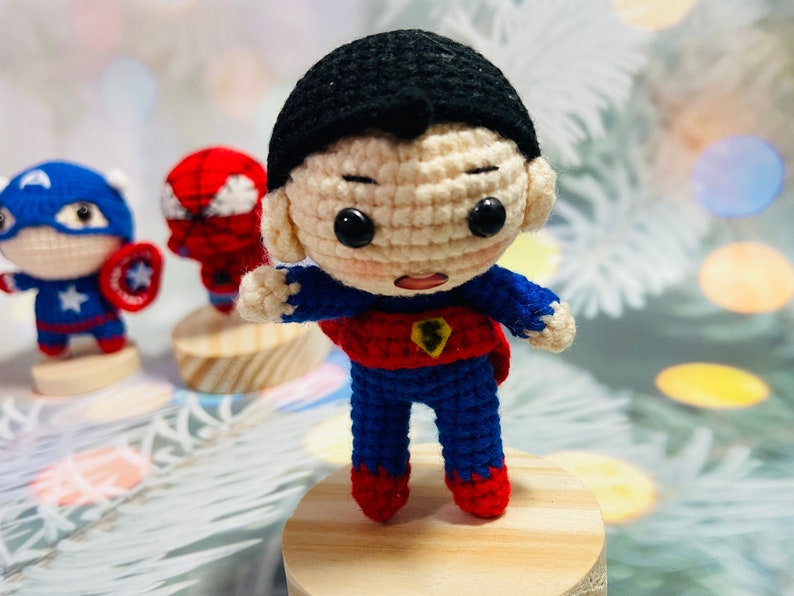 May include: A crocheted superhero doll with a red and blue costume, black hair, and a determined expression. The doll is standing on a wooden base.