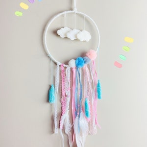 K&ouml;nnte beinhalten: Ein wei&szlig;er Traumf&auml;nger mit rosa, blauen und wei&szlig;en Pompons, wei&szlig;en Federn und wei&szlig;en Wolken, die am Reifen h&auml;ngen.