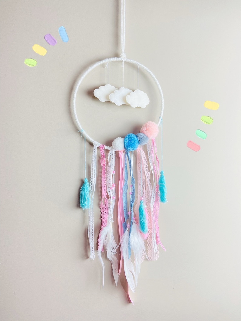 K&ouml;nnte beinhalten: Ein wei&szlig;er Traumf&auml;nger mit rosa, blauen und wei&szlig;en Pompons, wei&szlig;en Federn und wei&szlig;en Wolken, die am Reifen h&auml;ngen.
