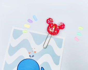May include: A red and clear plastic paper clip with a Mickey Mouse head design. The head is filled with red glitter and small gold stars. The clip is attached to a silver metal paper clip.