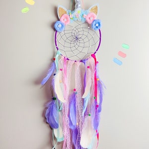 K&ouml;nnte beinhalten: Lilafarbener und wei&szlig;er Traumf&auml;nger mit einem Einhorn-Design. Der Traumf&auml;nger hat einen Perlenfransen mit Federn und Spitze. Das Einhorn-Design verf&uuml;gt &uuml;ber ein silbernes Horn, Glitzerohren und rosa und blaue Blumen.