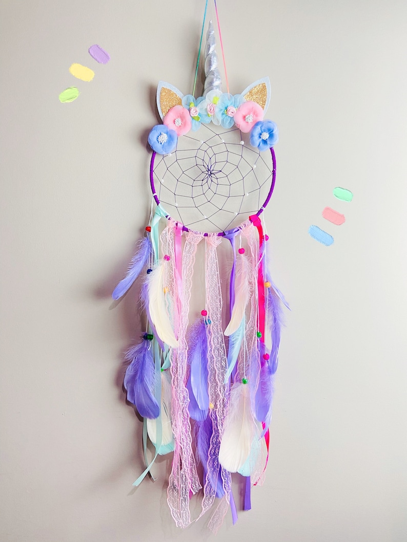 K&ouml;nnte beinhalten: Lilafarbener und wei&szlig;er Traumf&auml;nger mit einem Einhorn-Design. Der Traumf&auml;nger hat einen Perlenfransen mit Federn und Spitze. Das Einhorn-Design verf&uuml;gt &uuml;ber ein silbernes Horn, Glitzerohren und rosa und blaue Blumen.