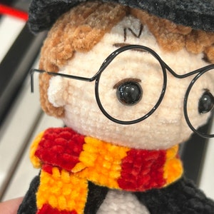 May include: A crocheted plush toy of a young boy wearing a black hat, round glasses, and a red and yellow scarf.
