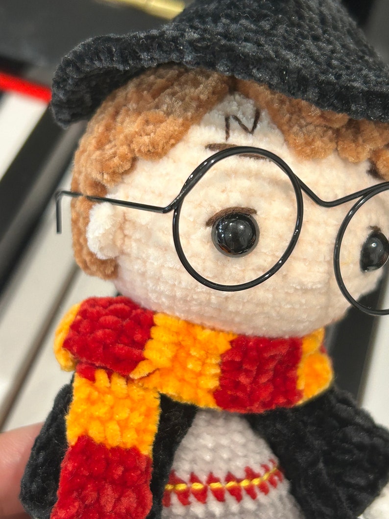 May include: A crocheted plush toy of a young boy wearing a black hat, round glasses, and a red and yellow scarf.