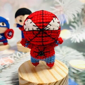 May include: A crocheted red and blue Spiderman figure with a black spider design on the chest. The figure is standing on a wooden base.