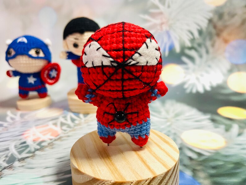 May include: A crocheted red and blue Spiderman figure with a black spider design on the chest. The figure is standing on a wooden base.