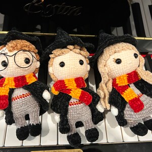 May include: Three crocheted Harry Potter characters, each wearing a black robe and a red and yellow scarf. The characters are standing on a white and black keyboard.