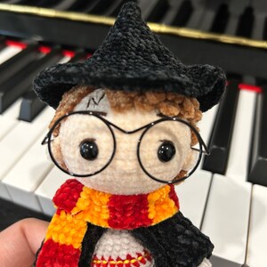 May include: A crocheted Harry Potter doll wearing a black hat, round glasses, a red and yellow scarf, and a black robe.