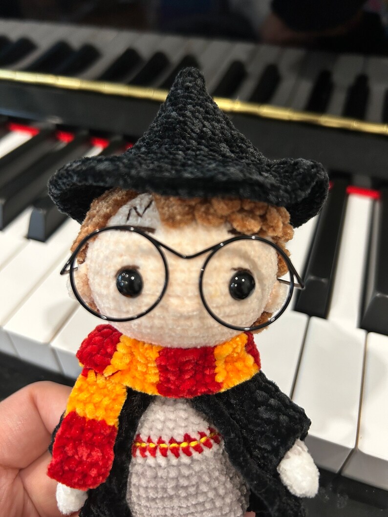 May include: A crocheted Harry Potter doll wearing a black hat, round glasses, a red and yellow scarf, and a black robe.