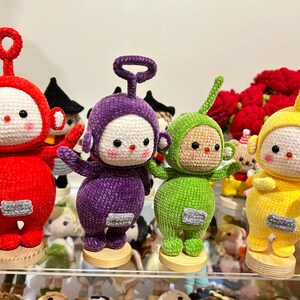 May include: Five crocheted plush toys in the shape of characters from the children's television show Teletubbies. The toys are red, purple, green, and yellow. They are standing on wooden bases.