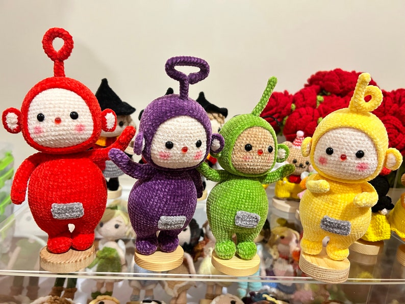 May include: Five crocheted plush toys in the shape of characters from the children's television show Teletubbies. The toys are red, purple, green, and yellow. They are standing on wooden bases.