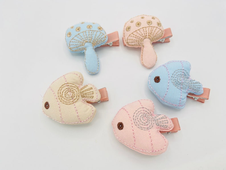 May include: Set of five fabric hair clips featuring embroidered designs of mushrooms and fish. The clips are in shades of blue, pink, and beige.