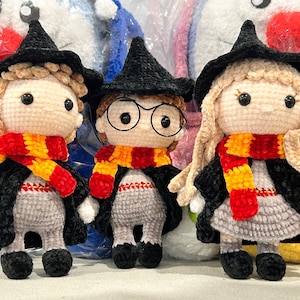 May include: Three crocheted plush toys dressed as Harry Potter characters. The toys are wearing black robes, pointed hats, and scarves with red and yellow stripes.