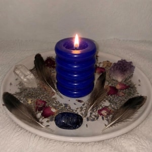 May include: A lit, ribbed, dark blue pillar candle sits on a white plate surrounded by scattered dried lavender, small pink rose petals, four brown and white feathers, a clear quartz crystal, an amethyst cluster, and a smooth dark blue stone.