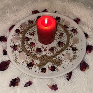 May include: A white circular plate holds a lit red pillar candle in the center, surrounded by a circular jute rope with a knot at the bottom. Scattered around the candle are dried dark red rose petals, small green herbs, and pale pink crystals.