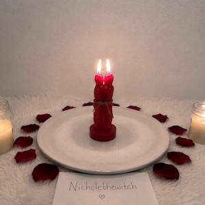 May include: A red figural candle shaped like two figures embracing, tied with twine, and lit with two flames. The candle sits on a white plate surrounded by red rose petals and two smaller white candles. A card reads Nicholethewitch with a heart.